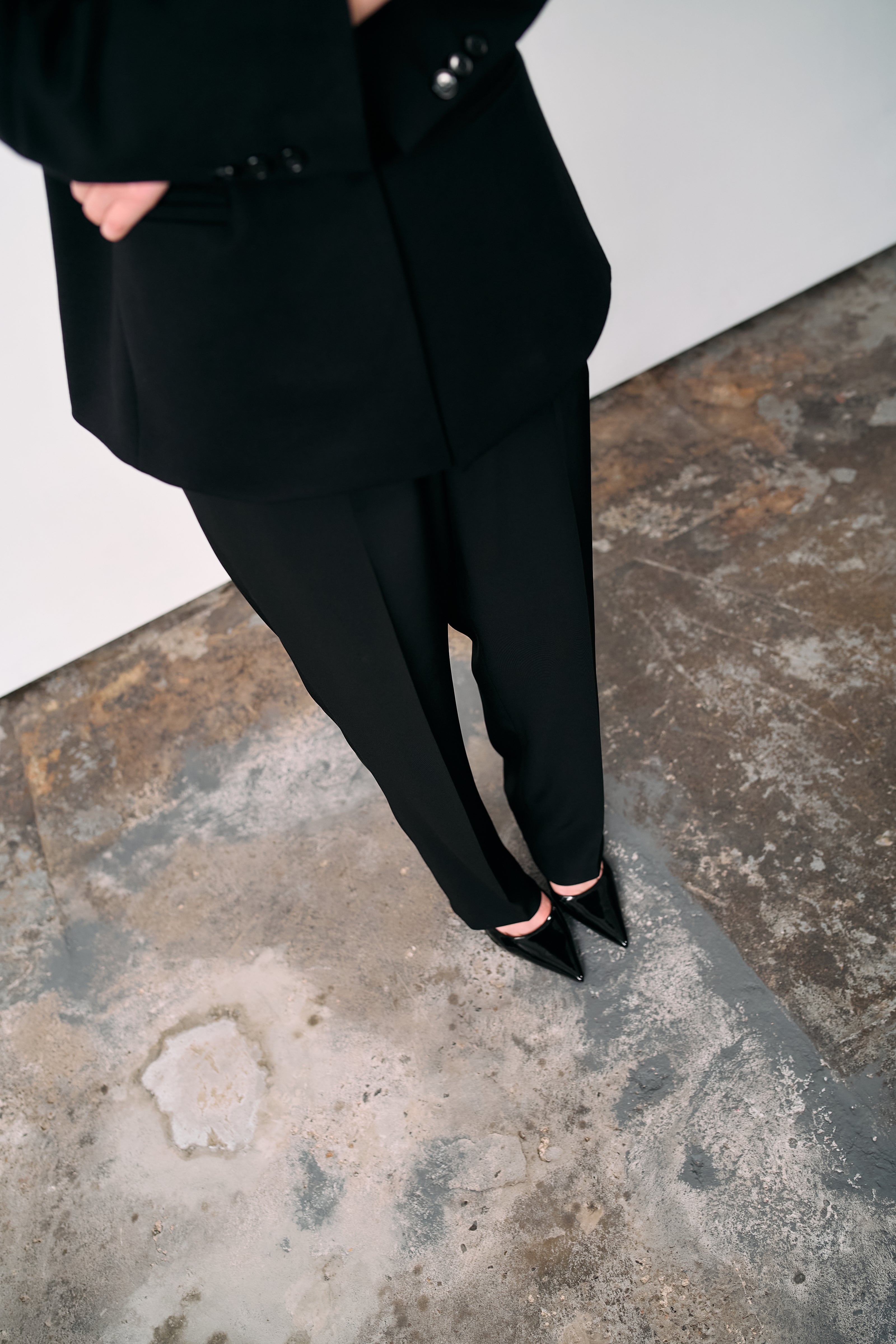 Black tailored trousers styled with a structured blazer and pointed heels, photographed from above on a textured concrete floor in a minimalist studio.