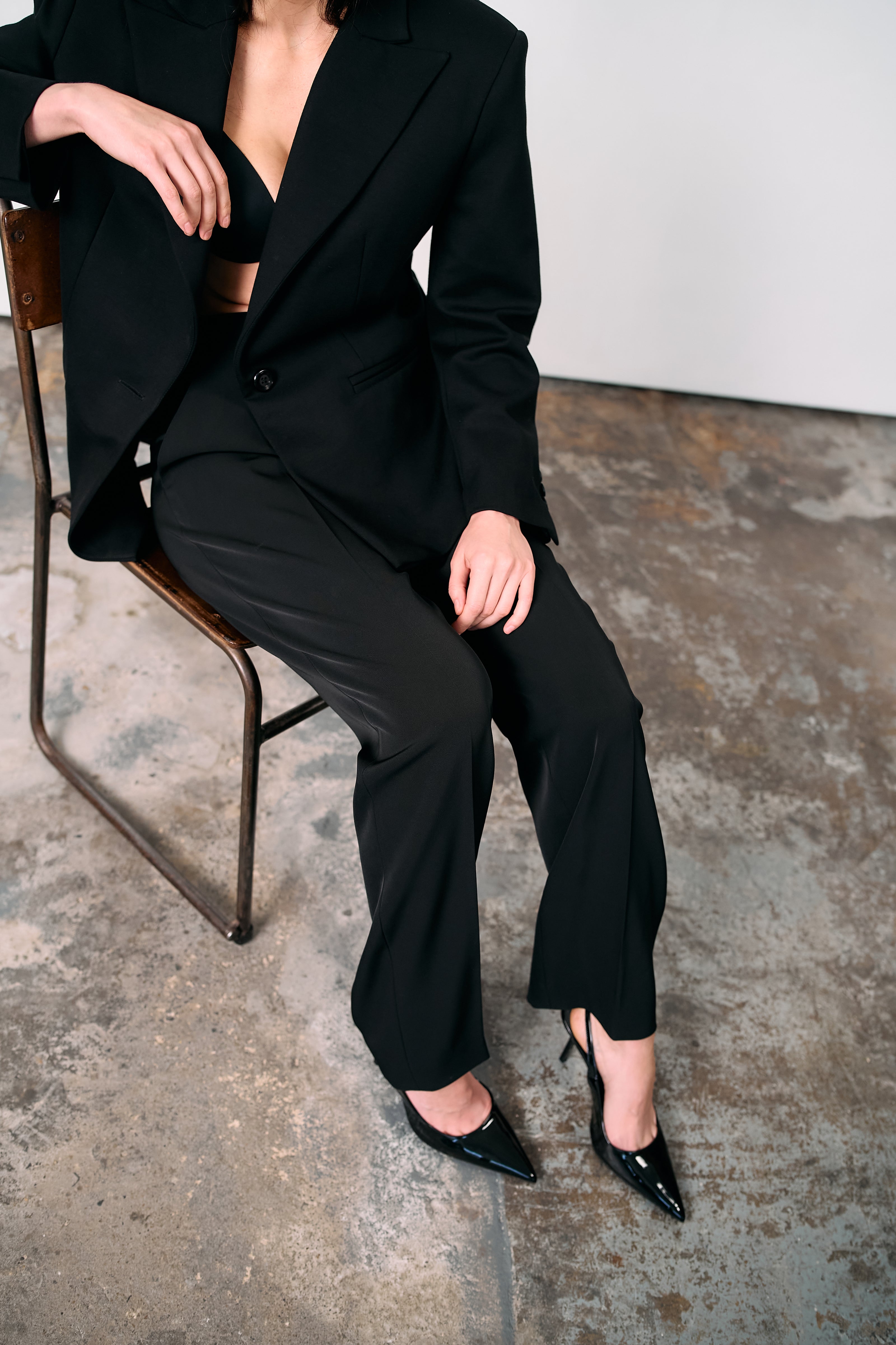 Black high-waisted tailored trousers styled with a structured blazer, worn open over a bralette, photographed seated on a metal chair against a concrete floor in a minimalist studio.