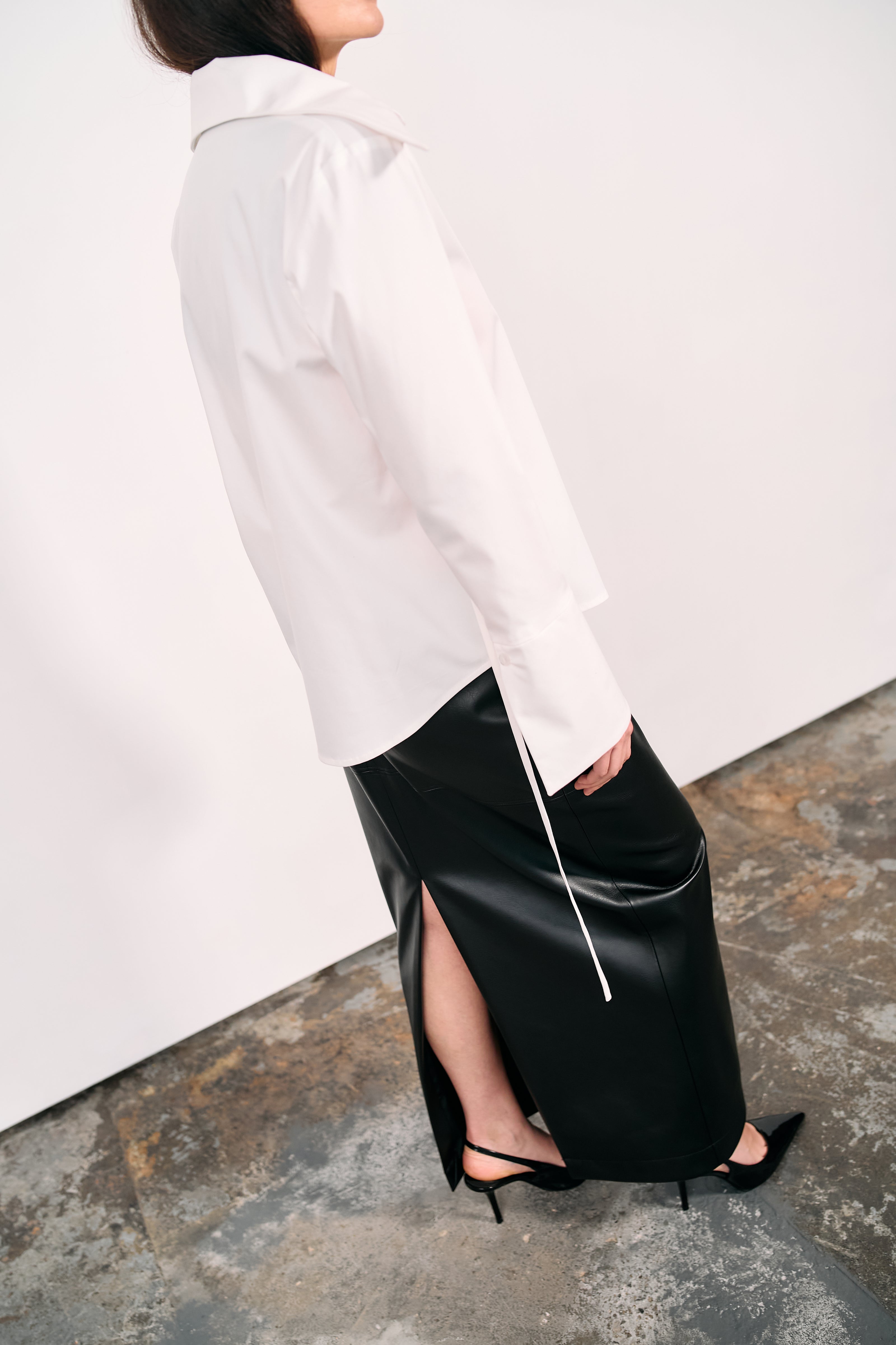 Black high-waisted PU maxi skirt with a side slit, styled with pointed heels and a crisp white shirt, photographed in a minimalist studio with a concrete floor and white backdrop.