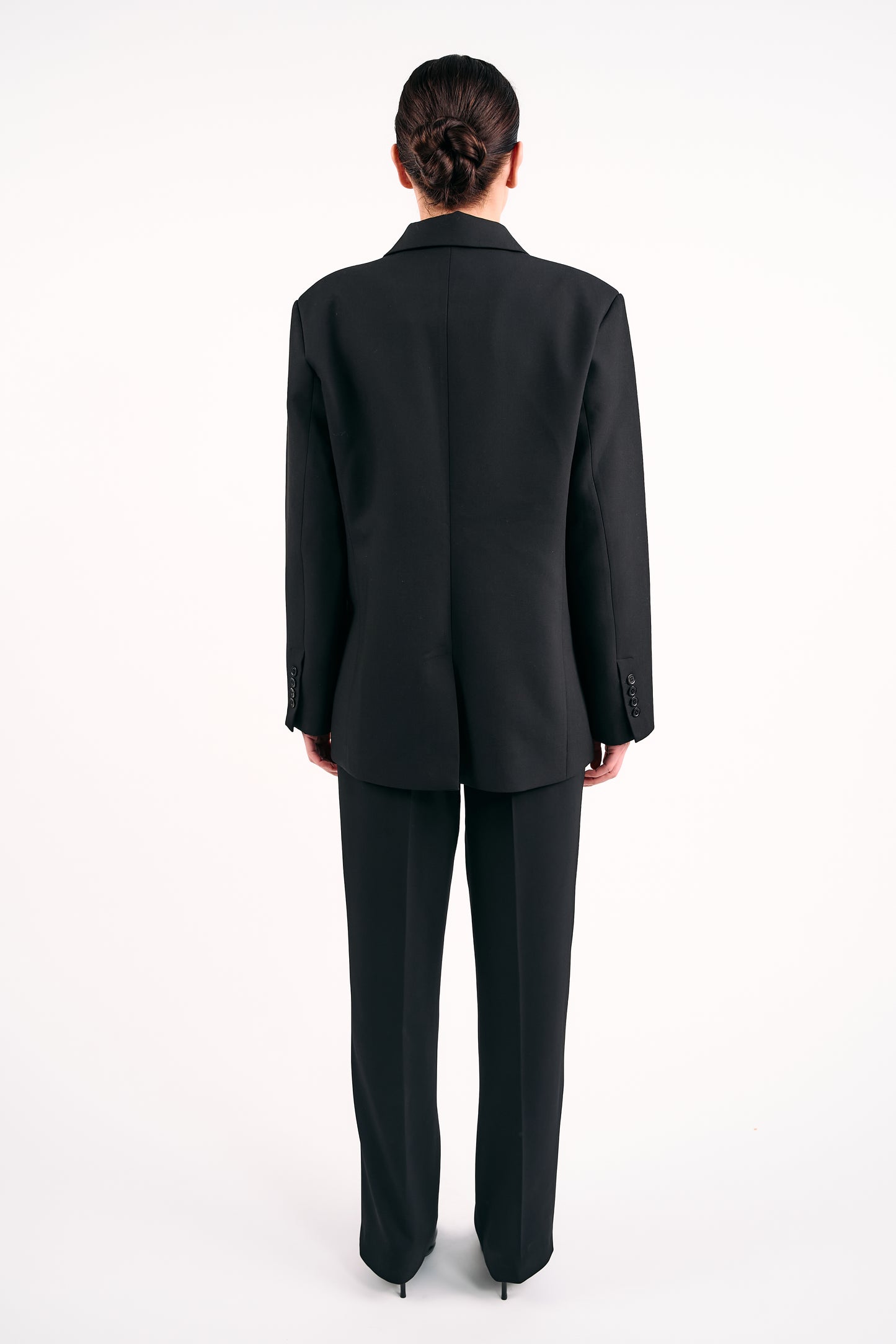 Woman wearing a black tailored blazer and matching black trousers, photographed from the back against a white background.