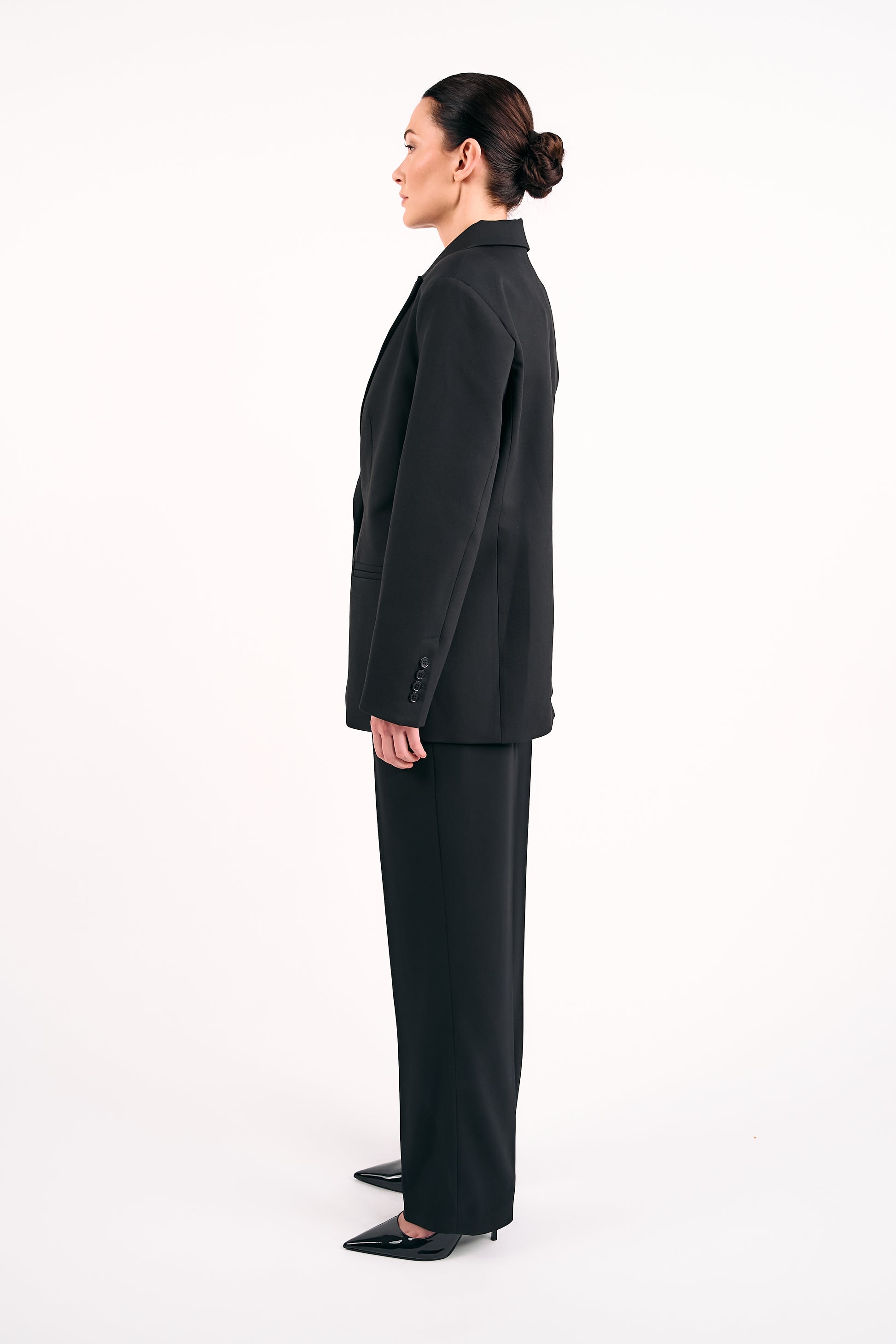 Woman wearing a black tailored blazer and matching black trousers, standing in profile against a white background.