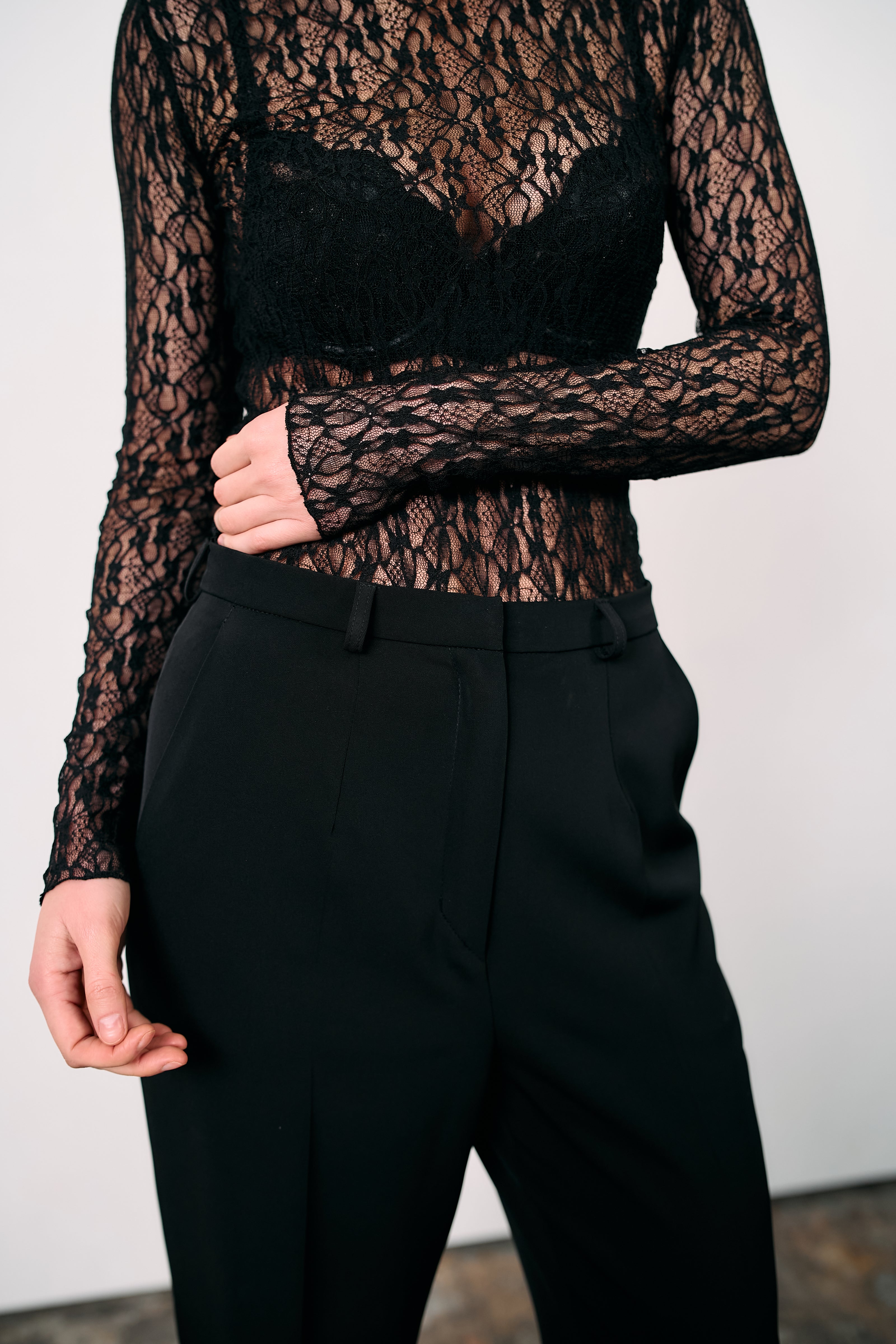 Black high-waisted tailored trousers styled with a sheer black lace top, photographed against a minimalist white background.