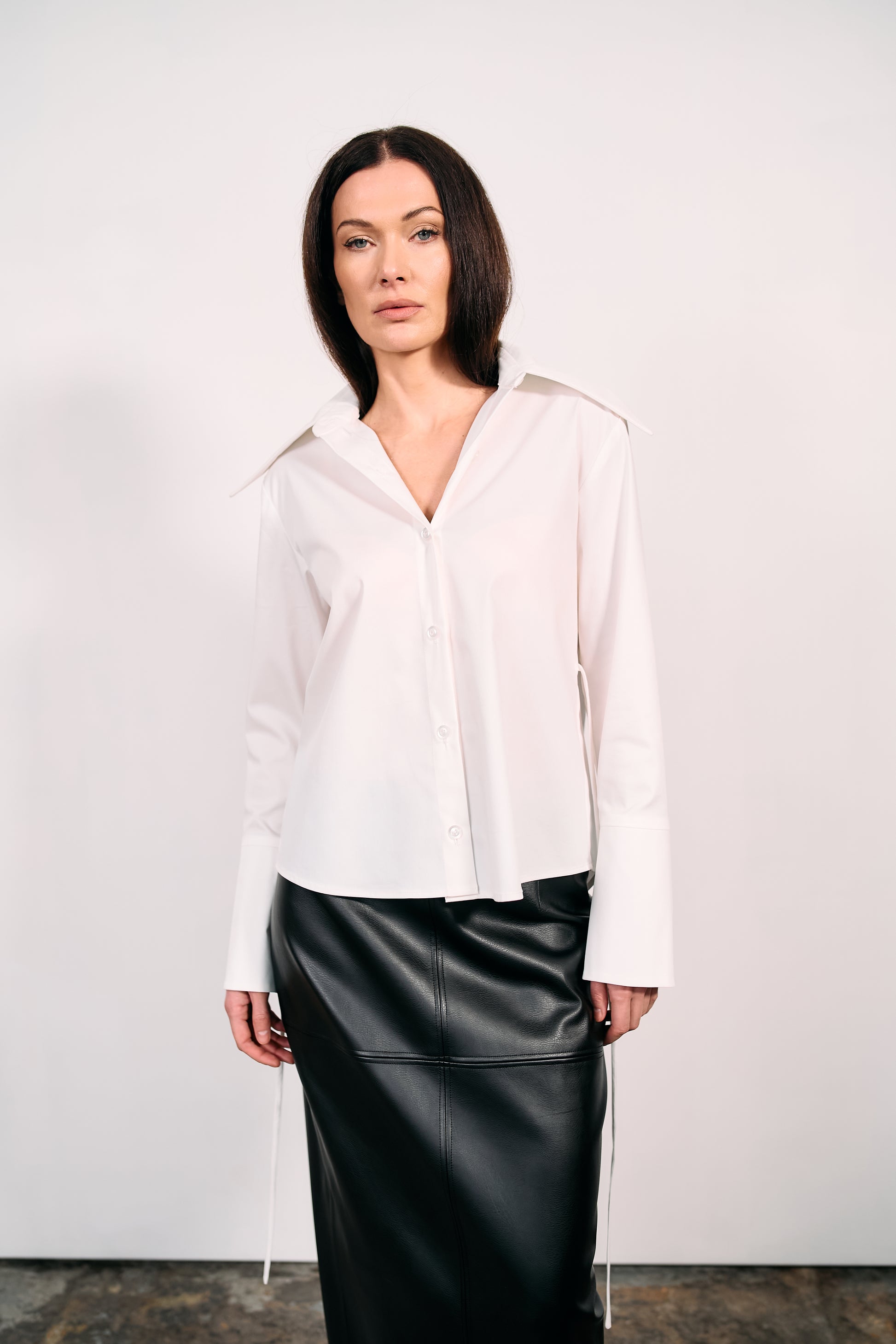 Woman wearing a white shirt and black PU leather maxi skirt, standing against a white wall and concrete studio floor.