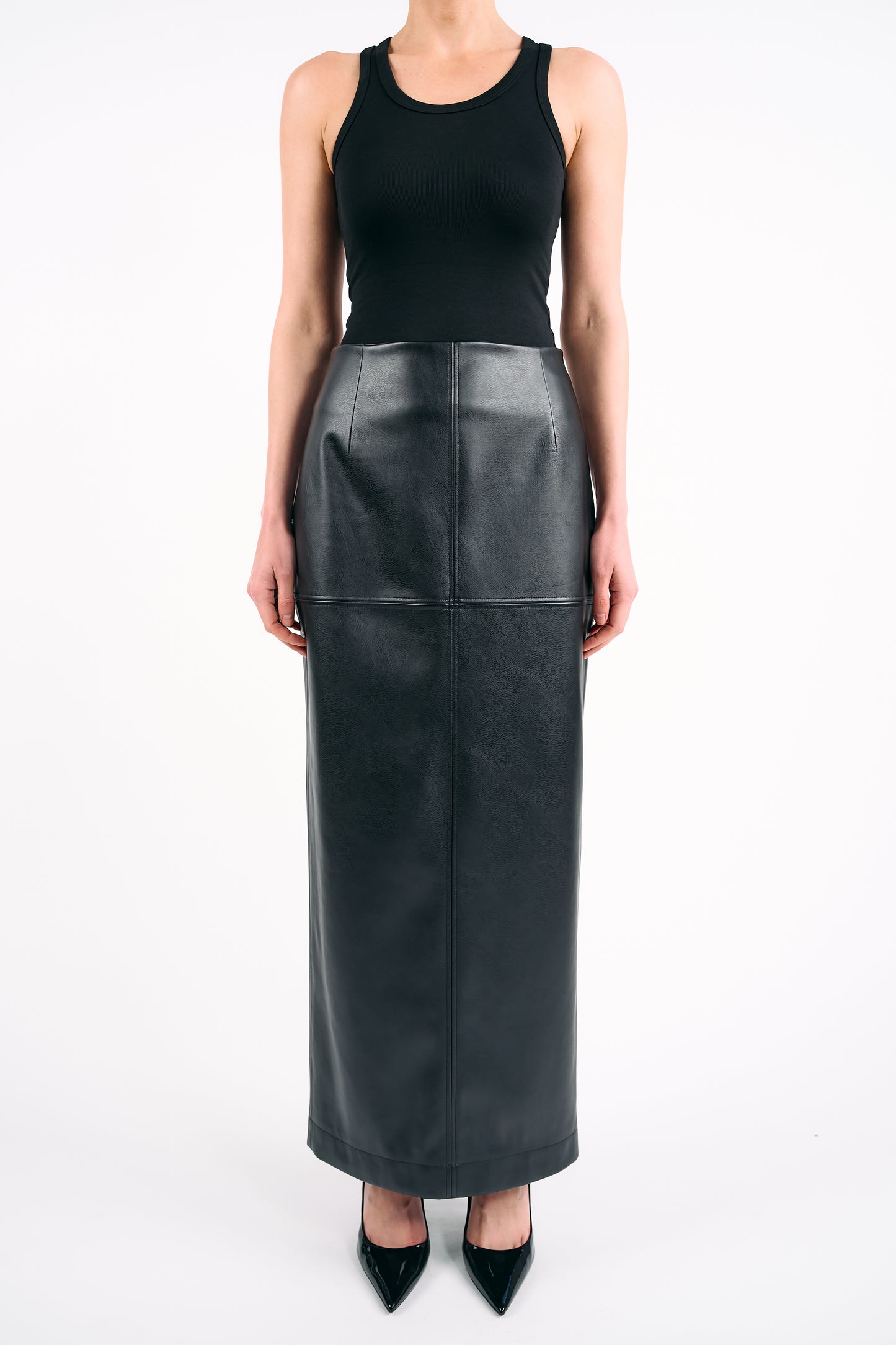 Black high-waisted PU maxi skirt styled with a fitted black tank top and pointed heels, photographed against a white background in a minimalist studio setting.