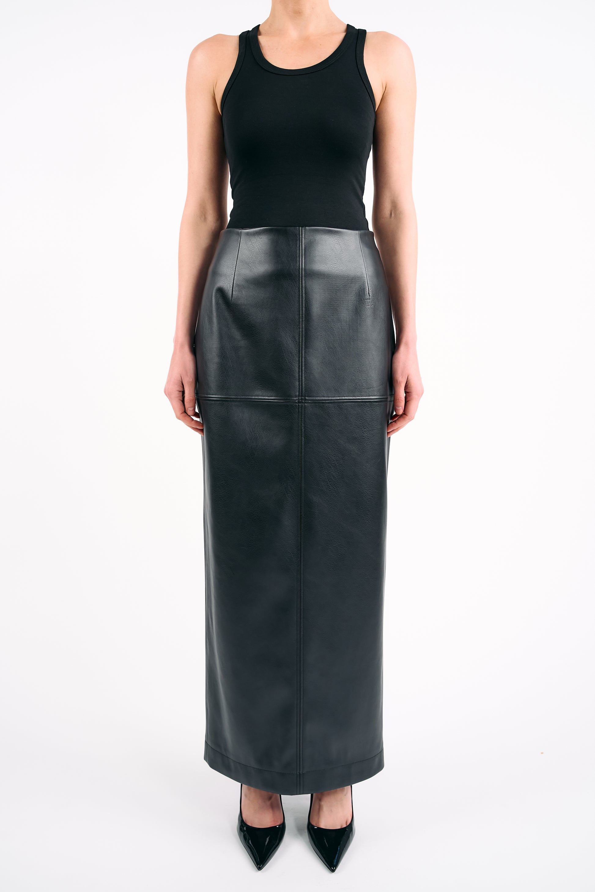 Black high-waisted PU maxi skirt styled with a fitted black tank top and pointed heels, photographed against a white background in a minimalist studio setting.
