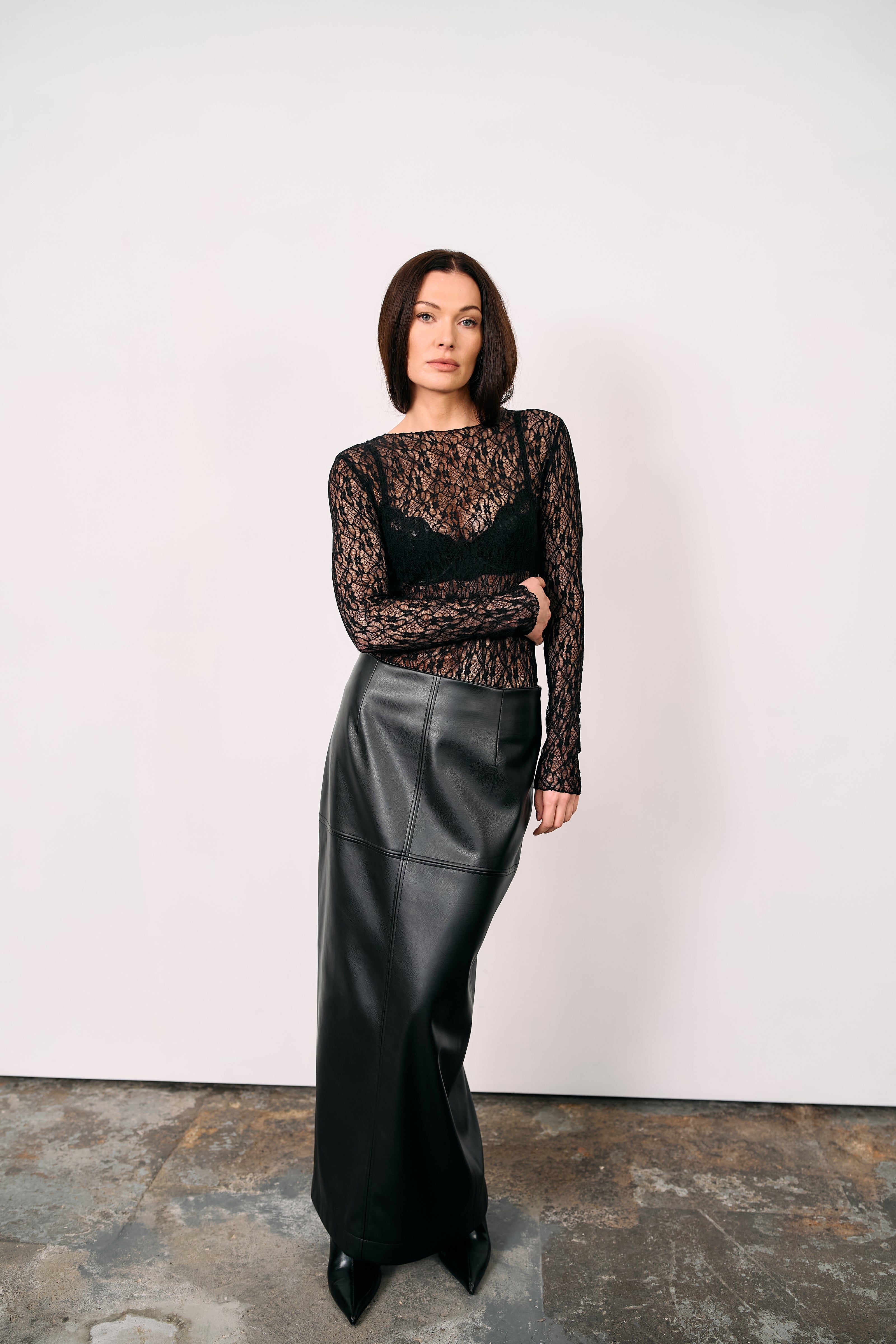 Black high-waisted leather maxi skirt styled with a sheer lace top and pointed boots, photographed in a minimalist studio against a white background.