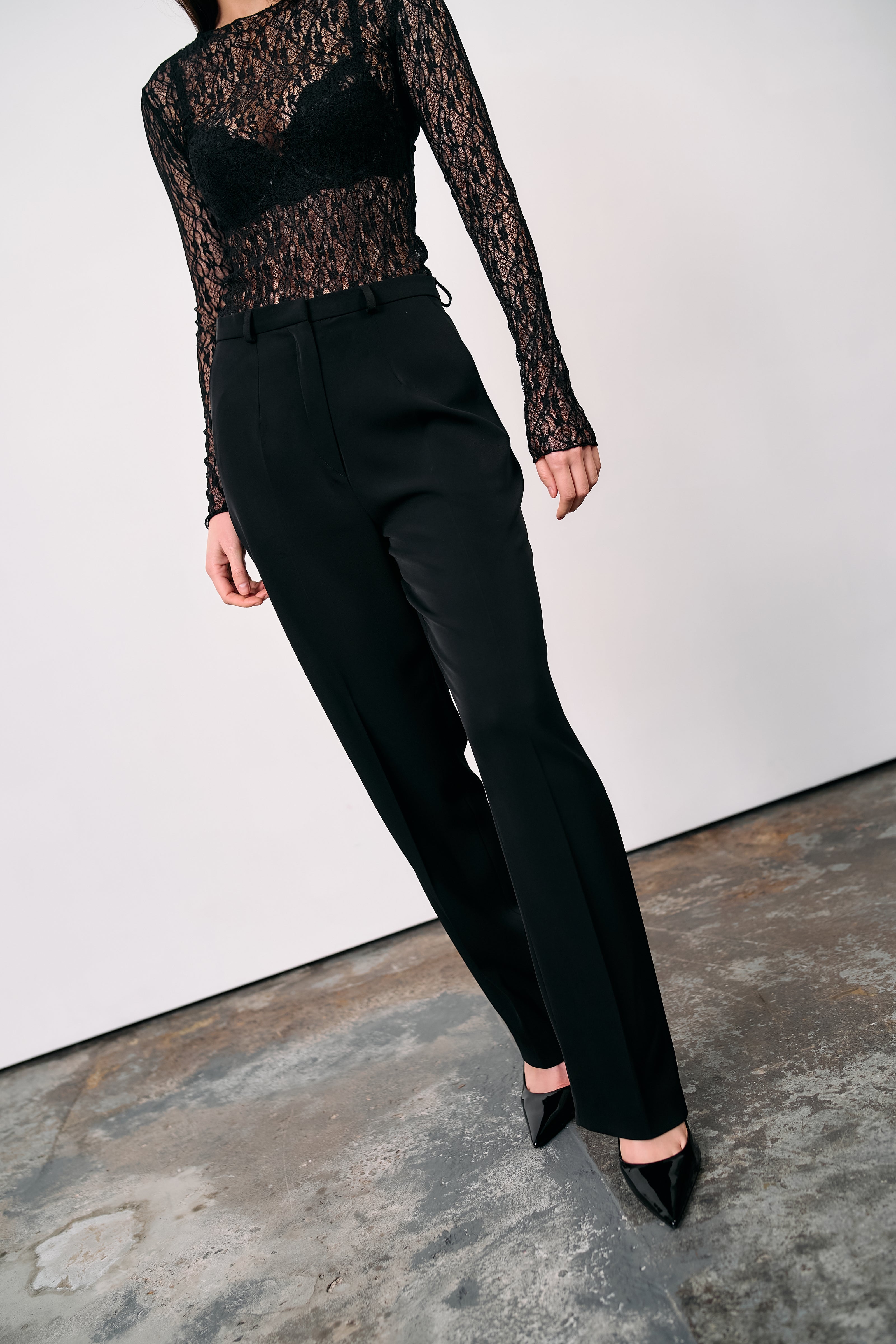 Black high-waisted tailored trousers styled with a sheer lace top and pointed heels, photographed in a minimalist studio on a concrete floor.