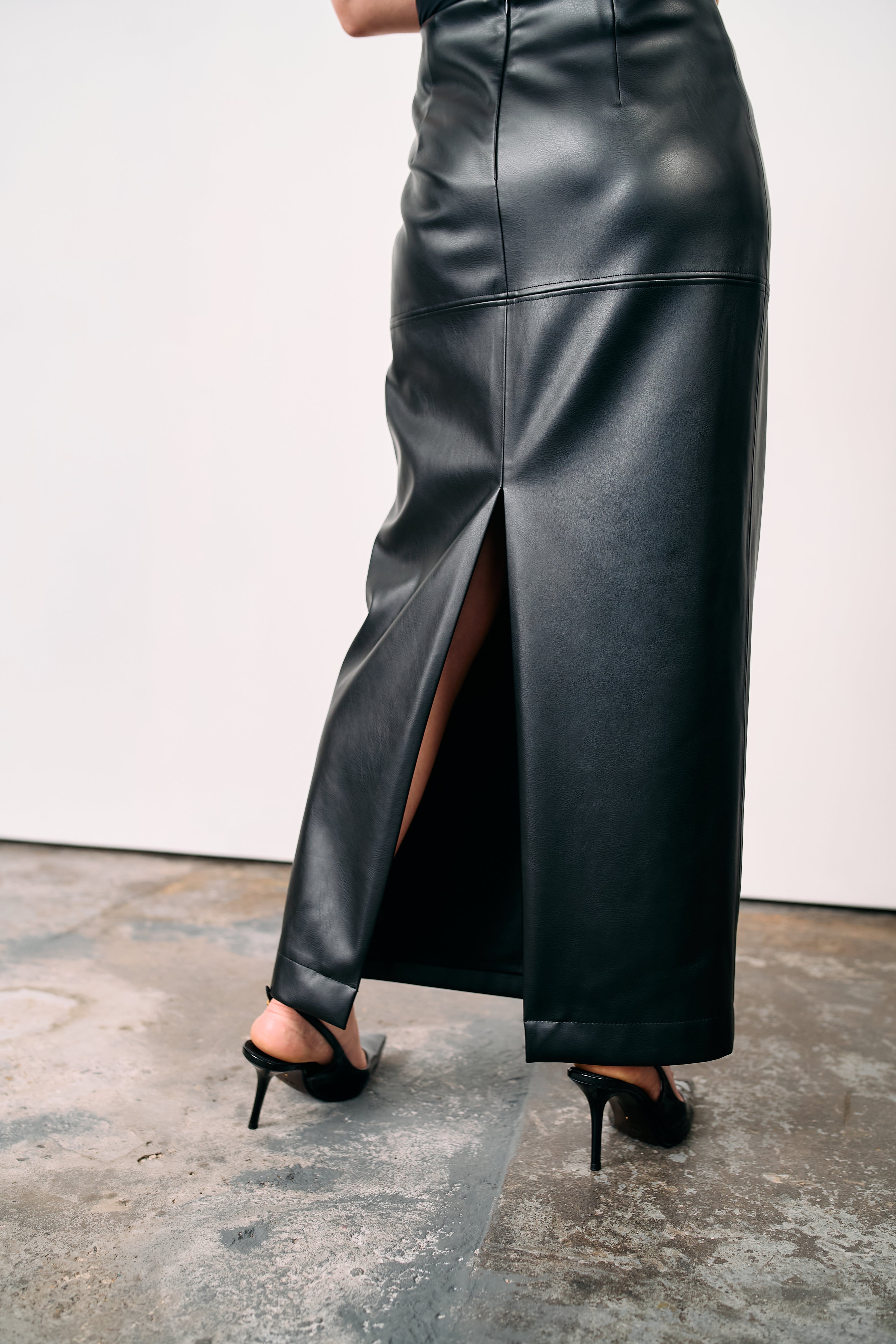 Black high-waisted leather maxi skirt with back slit, styled with lace top and heeled boots, photographed on a concrete floor in a minimalist studio.