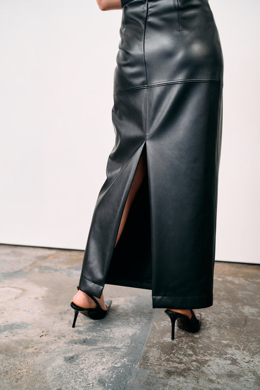Black high-waisted leather maxi skirt with back slit, styled with lace top and heeled boots, photographed on a concrete floor in a minimalist studio.