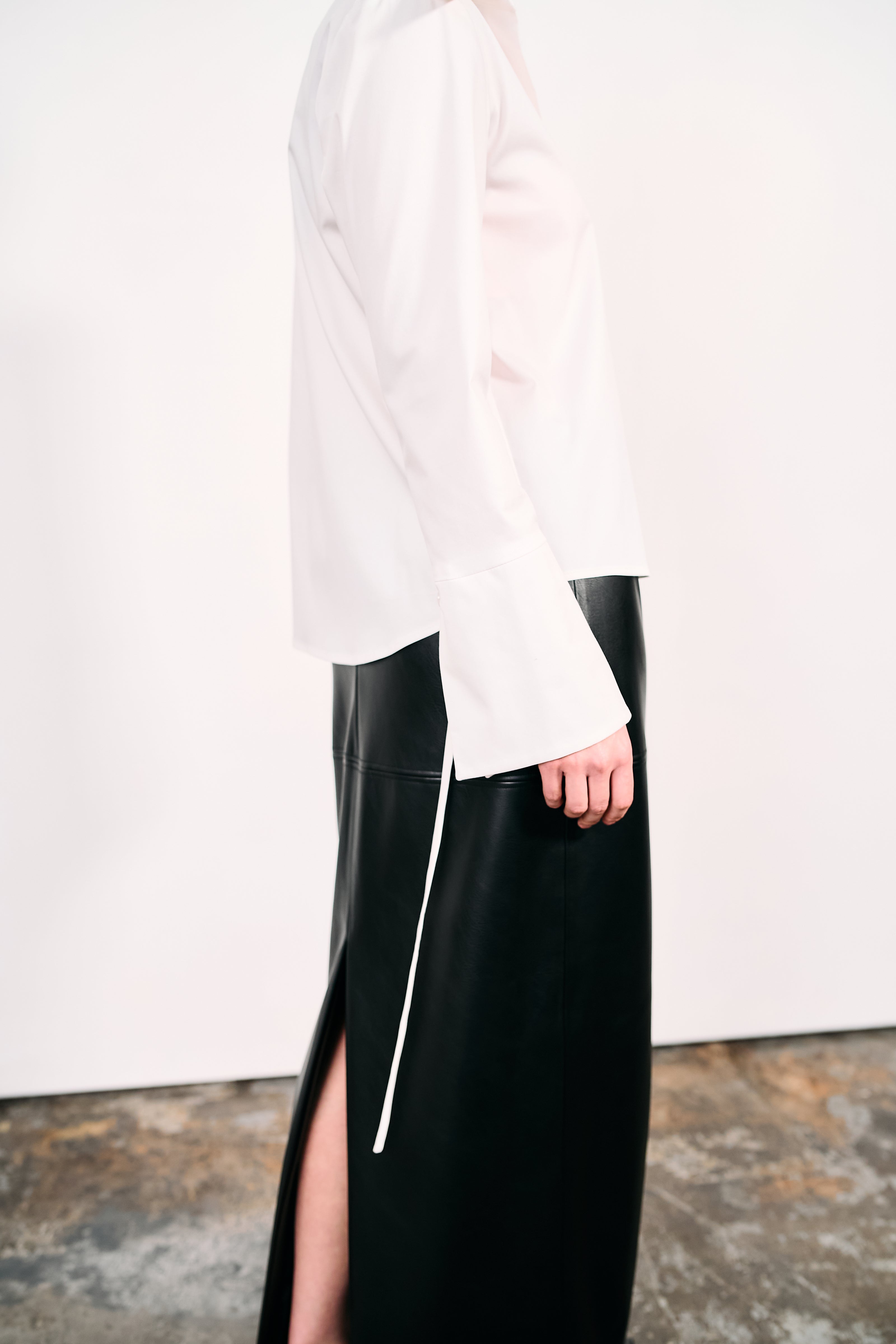 Woman wearing a white shirt and black PU leather maxi skirt, photographed against a white wall and concrete studio floor.