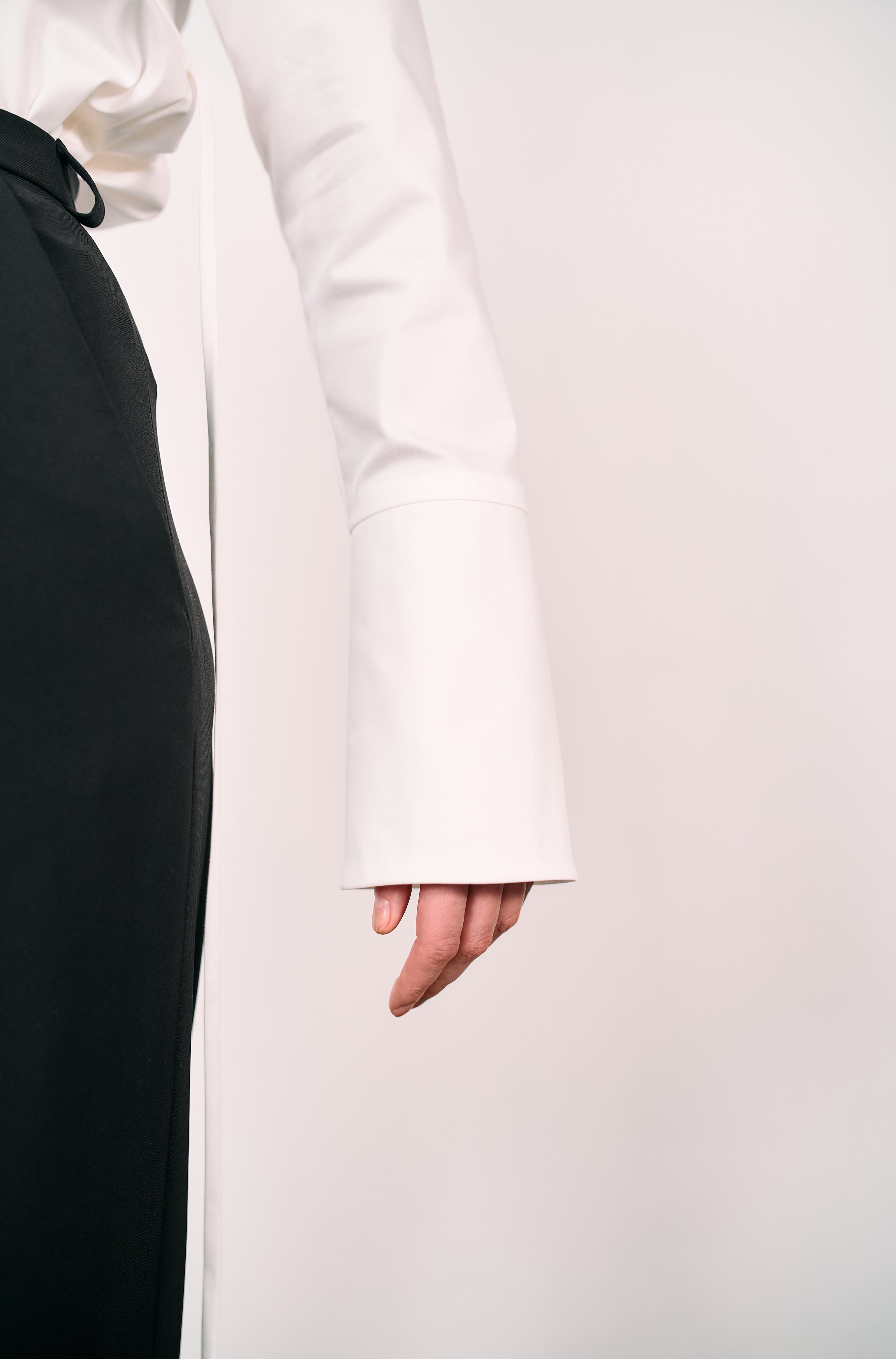 Detail shot of a white long-sleeve shirt with flared cuff paired with black tailored trousers, photographed in a minimalist studio setting.