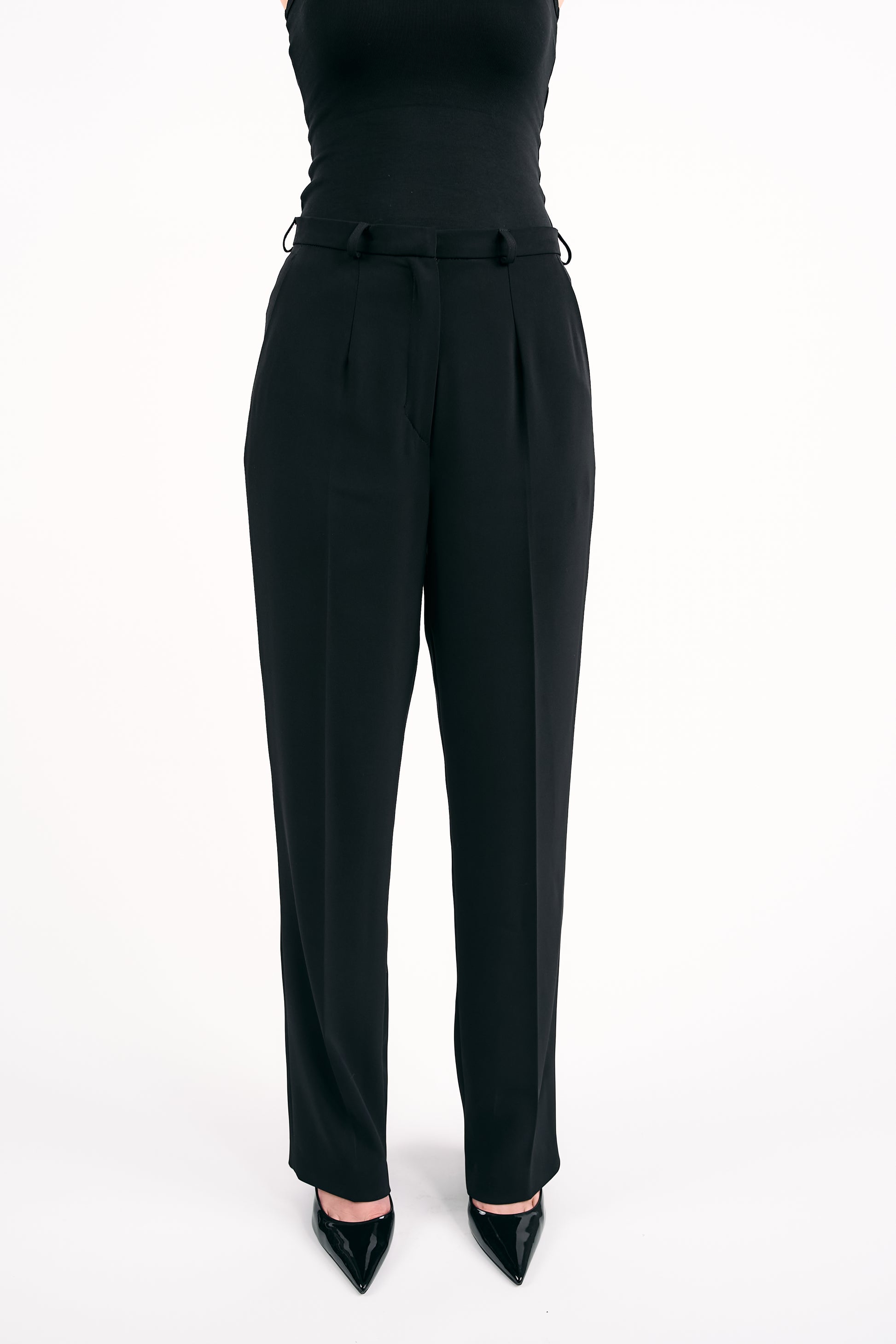 Woman wearing black high-waisted trousers with a belt and a fitted black sleeveless top against a white background.