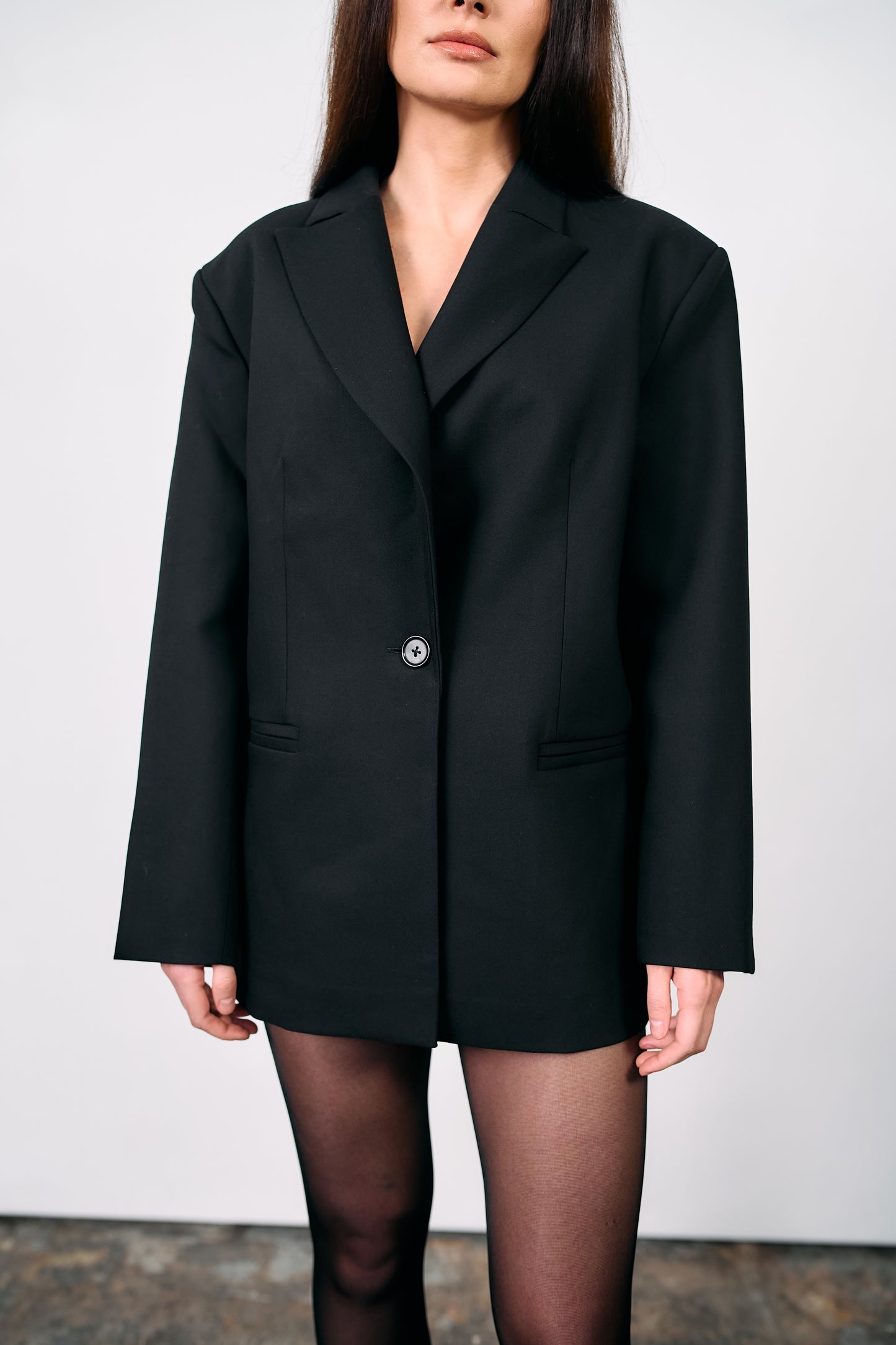 Woman wearing an oversized black tailored blazer styled as a dress, paired with pointed heels, photographed against a white wall on a concrete studio floor.