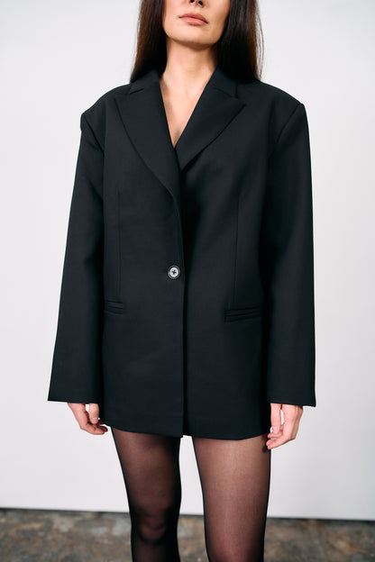 Woman wearing an oversized black tailored blazer styled as a dress, paired with pointed heels, photographed against a white wall on a concrete studio floor.