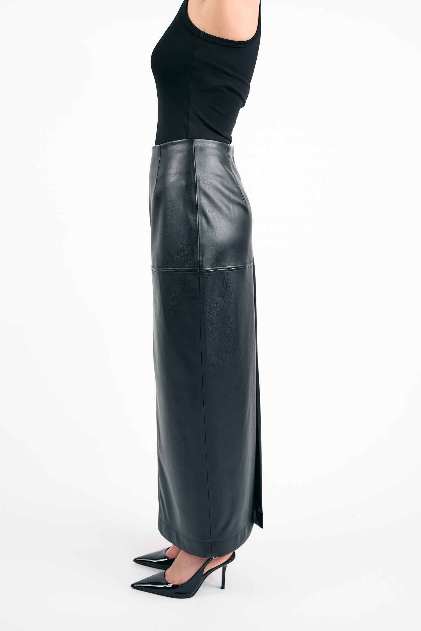 Woman wearing a black PU leather maxi skirt with a side slit and a black sleeveless top, photographed from the side against a white background.