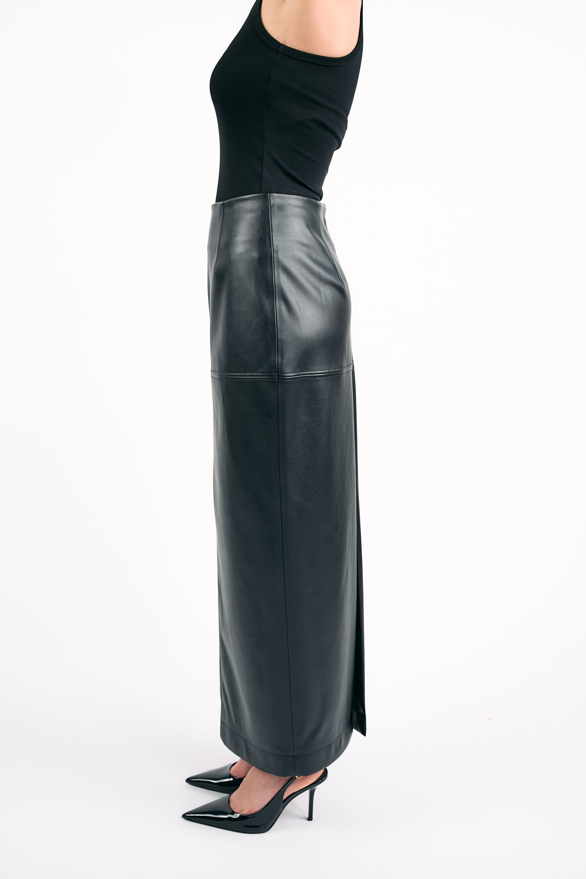 Woman wearing a black PU leather maxi skirt with a side slit and a black sleeveless top, photographed from the side against a white background.