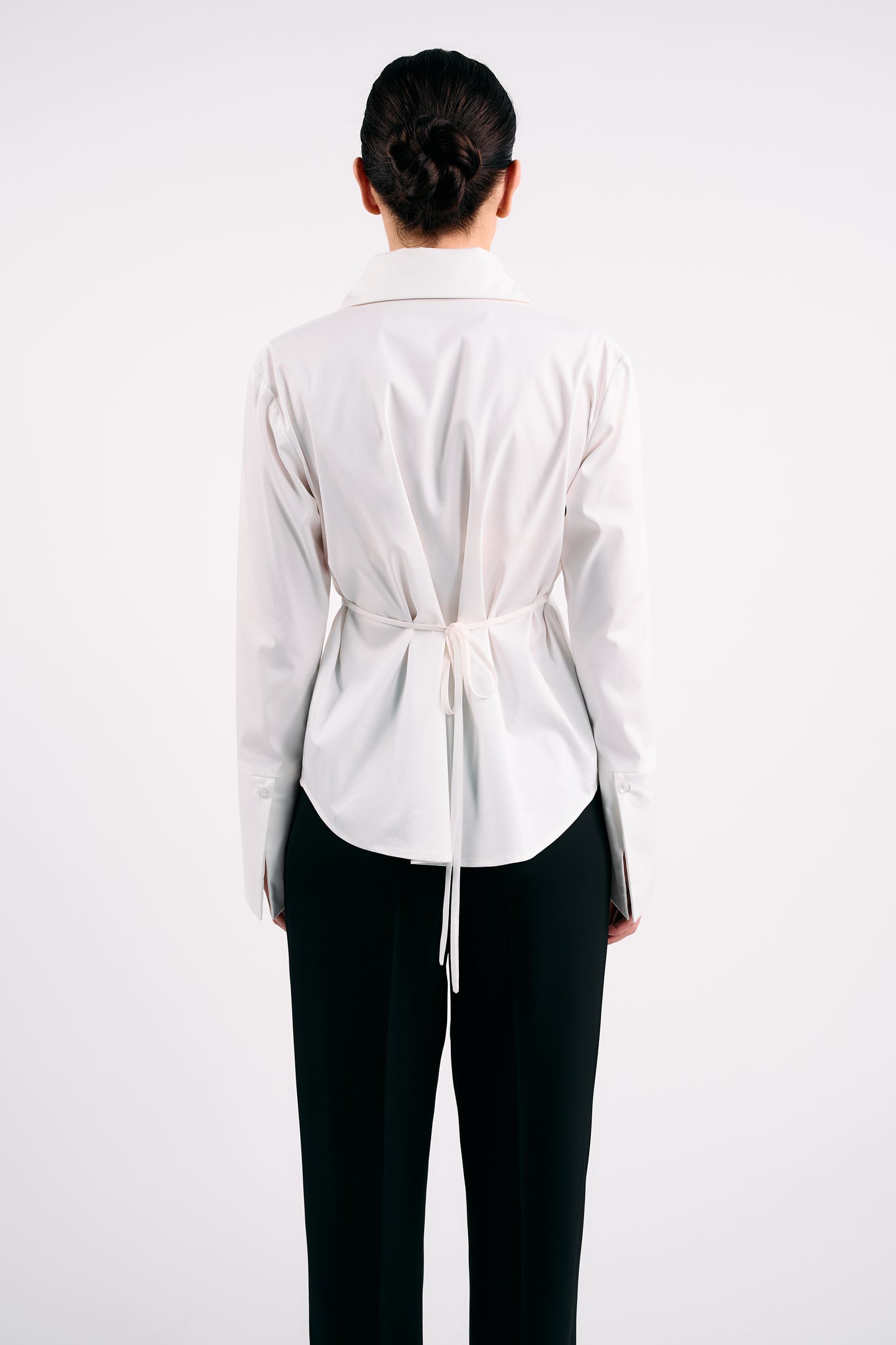 White structured shirt with back tie detail, styled with black tailored trousers, photographed from behind against a plain white background.