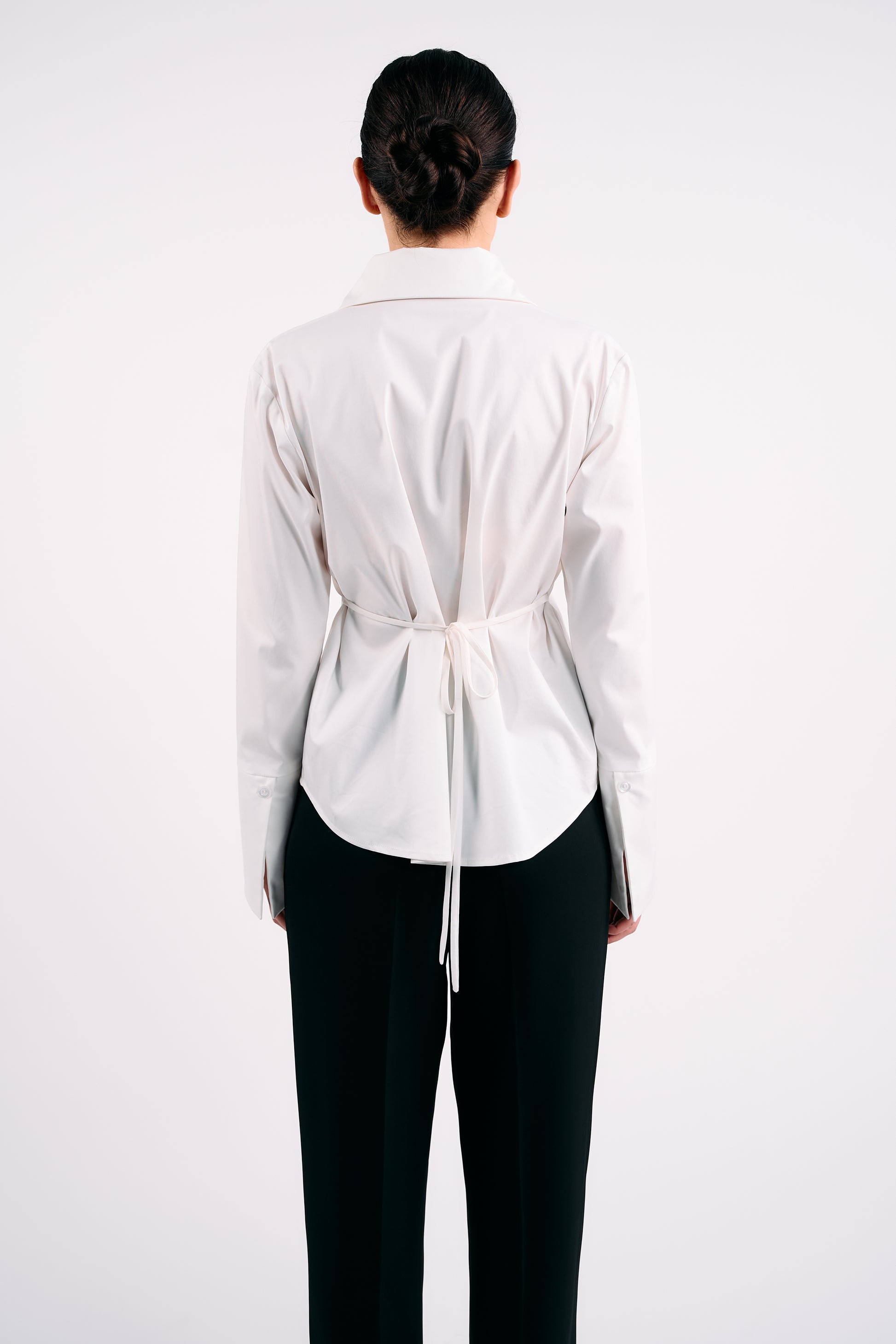 White structured shirt with back tie detail, styled with black tailored trousers, photographed from behind against a plain white background.