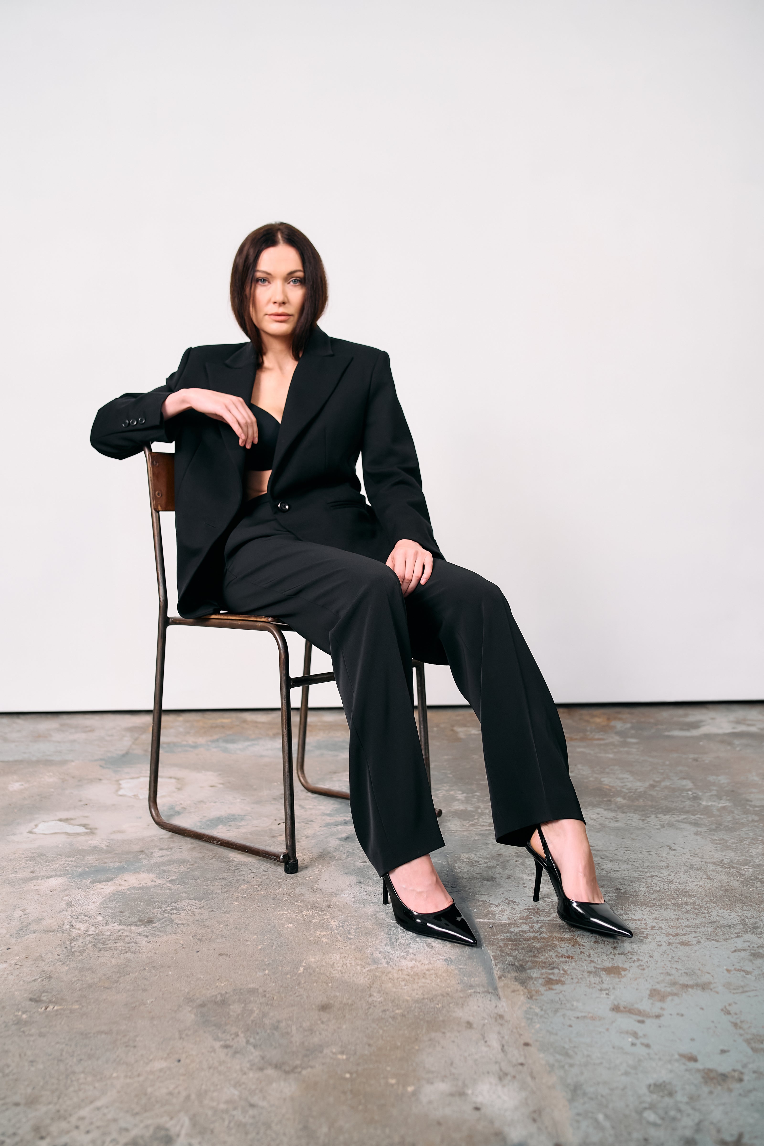Black high-waisted tailored trousers styled with a structured blazer, worn open over a bralette, photographed seated on a metal chair against a concrete floor in a minimalist studio.