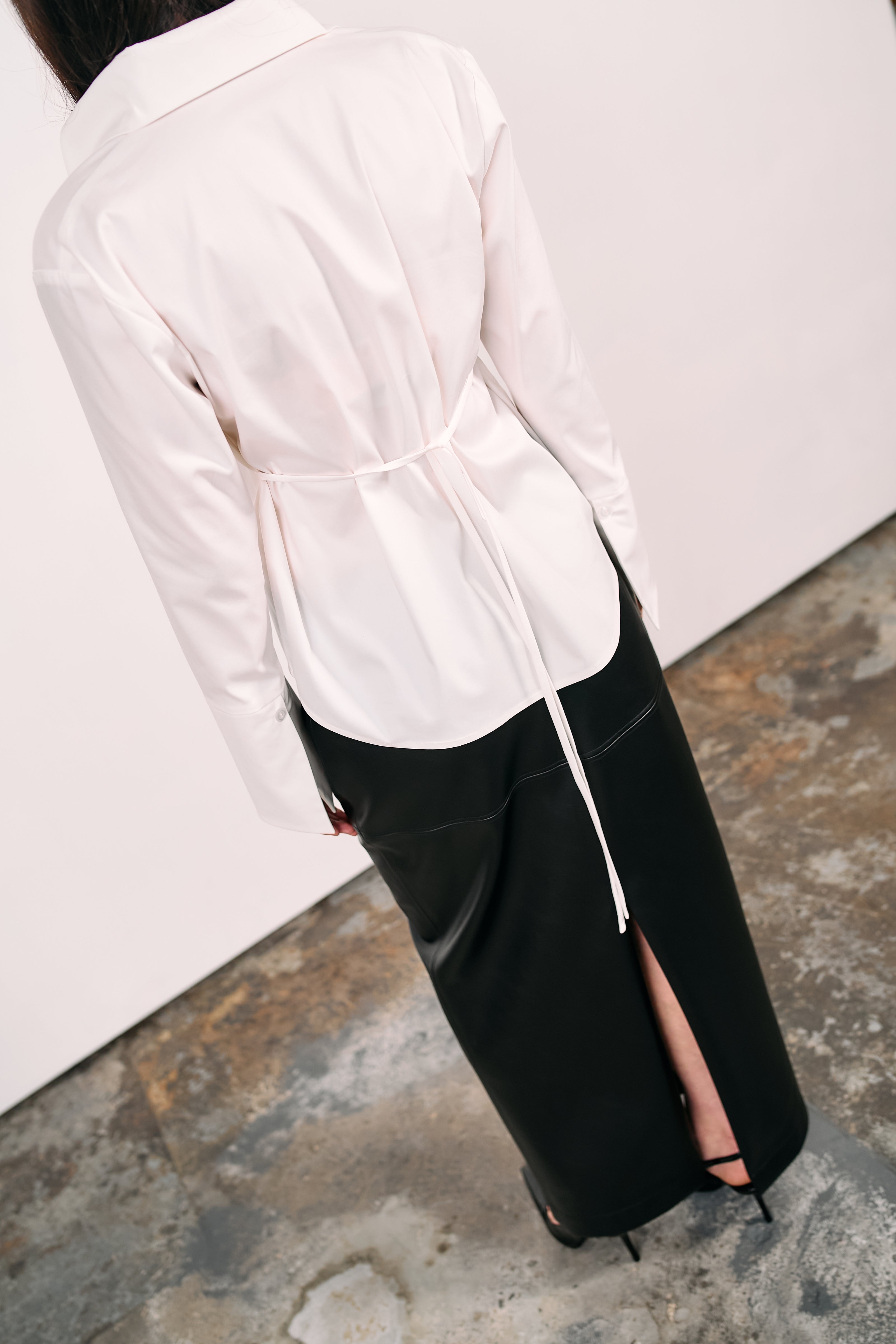 Woman wearing a white blouse with a back tie detail, paired with a black PU maxi skirt featuring a back slit, photographed against a white wall on a concrete floor.