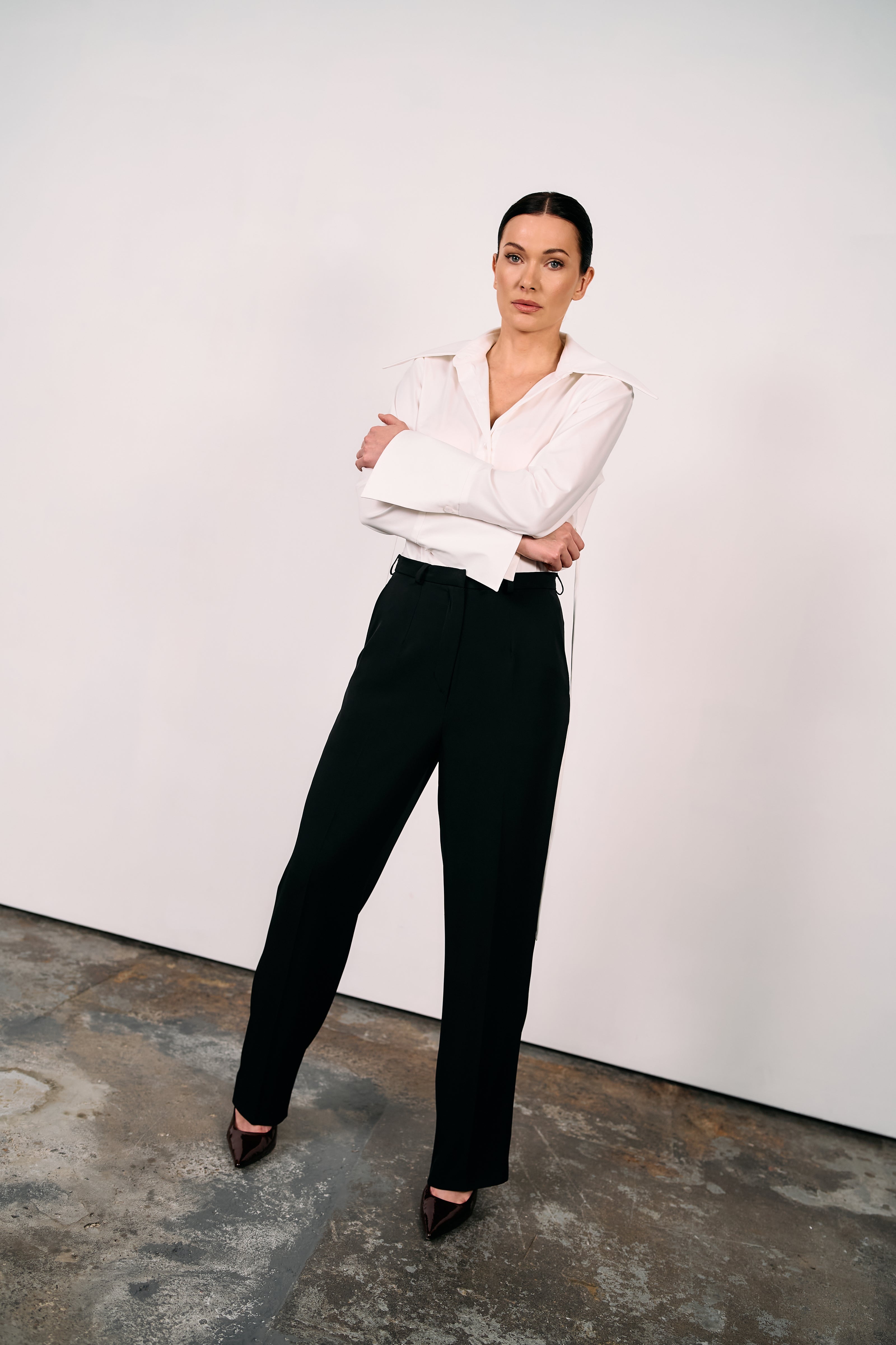 Woman wearing black wide-leg tailored trousers and a crisp white shirt, standing in a minimalist studio space.